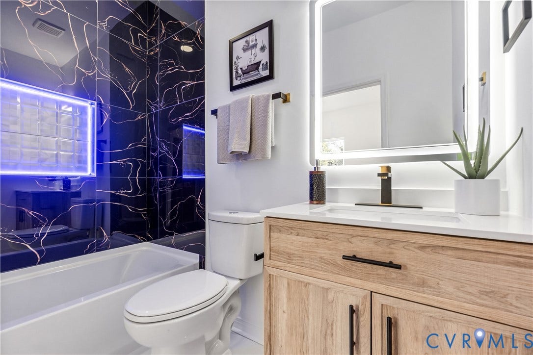 223 S Crater Road, Petersburg, VA 23803 Bathroom featuring wood-finish cabinetry, a rectangular undermount sink with matte black hardware, a backlit wall-mounted mirror, and black wall tile with copper-toned veining in the shower area