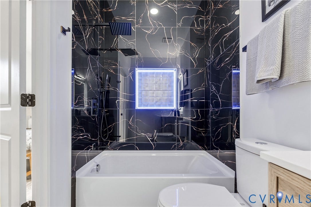 223 S Crater Road, Petersburg, VA 23803 Bathroom featuring black vein-patterned wall tile, square rain-style shower head, integrated LED-lit glass block window, white soaking tub, and vanity with stone countertop