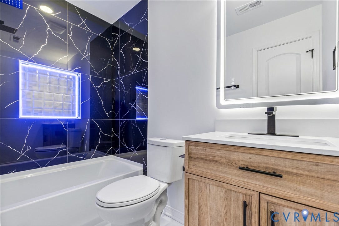 223 S Crater Road, Petersburg, VA 23803 Bathroom featuring black tile with white veining, wood-finish vanity with white countertop, recessed glass block niche, and integrated LED-backlit mirror