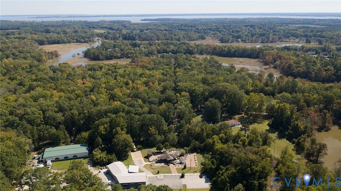 234 Sandy Bay Road, Williamsburg, VA 23185 Aerial perspective of a sprawling property featuring multiple structures, paved hardscaping, a metal-roofed facility, and significant acreage with dense woodland and natural waterways