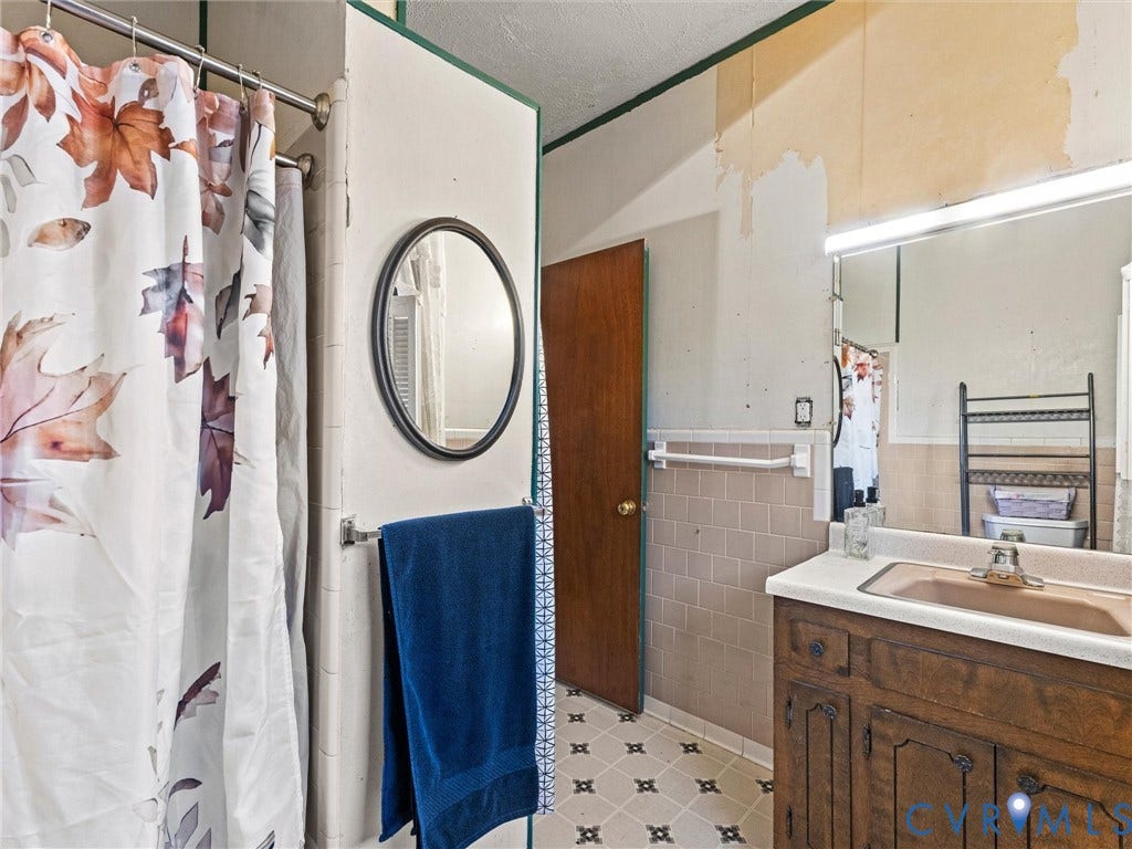 124 Suffolk Avenue, Chesterfield Highlands, Colonial Heights, VA 23834 Bathroom featuring a wood-finish vanity with an integrated sink, large wall mirror, circular accent mirror, tiled wall accents, shower, and patterned vinyl flooring