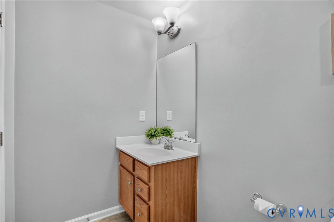 10851 White Dogwood Drive, Oakmont Villas, New Kent, VA 23140 Single-sink vanity with wood-finish cabinetry, solid-surface countertop, integrated basin, wall-mounted mirror, and dual-bulb fixture