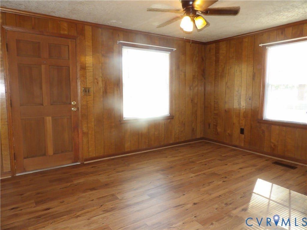 3206 South Street, Chester, VA 23831 Interior space featuring wood-panel wall treatments, wood-finish flooring, two large windows with horizontal blinds, a six-panel wood door, and a ceiling fan with light fixtures