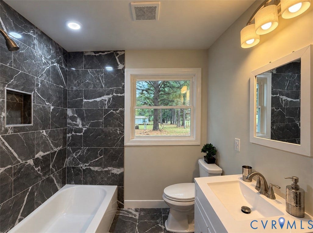 2800 Harris Hill Road, Dunnsville, VA 22454 Bathroom featuring a deep soaking tub with large-format dark stone-finish tile surround, integrated shower niche, white vanity with rectangular basin, and wall-mounted vanity mirror with integrated lighting