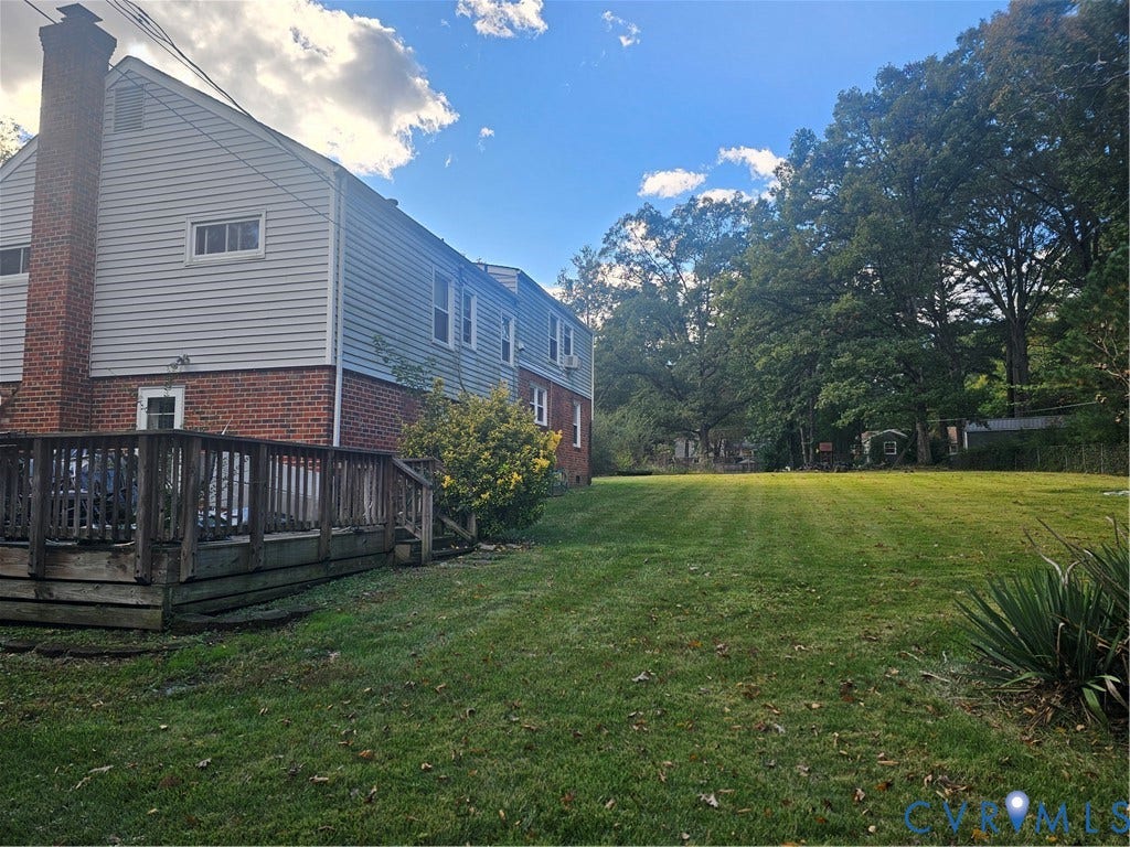 Rear exterior featuring a wood deck, red brick and horizontal siding faade, masonry chimney, and an expansive lawn with mature trees