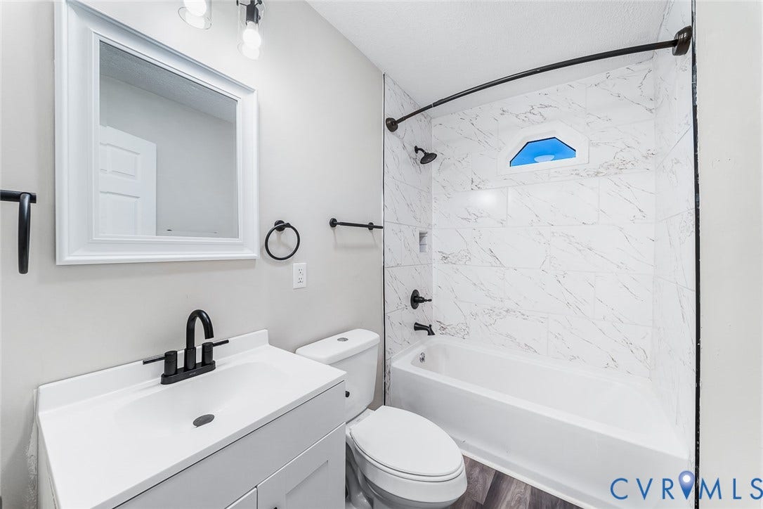 1802 Maury Street, Richmond, VA 23224 Bathroom featuring a white vanity cabinet, rectangular undermount sink with matte black faucet, wall-mounted rectangular mirror, and tub-shower surround with light-toned stone-finish tile and matte black hardware