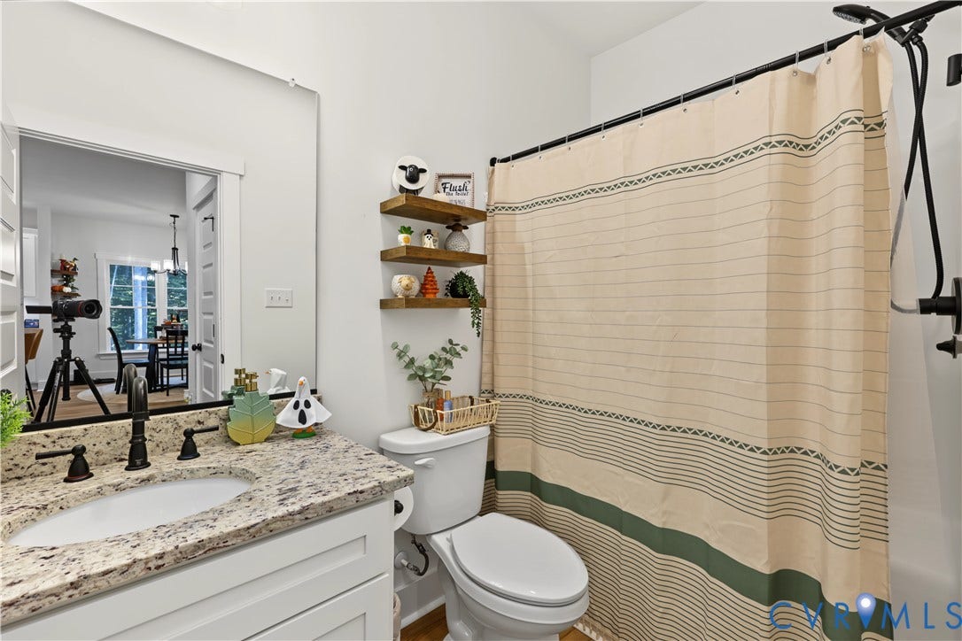 3851 County Line Road, Flemings Mill, Goochland, VA 23084 Bathroom featuring a stone-surface vanity with an integrated undermount sink, dark-finish faucet, wall-spanning mirror, three tiered floating wood shelves, and a tub-shower combination with a dark-finish rainfall showerhead