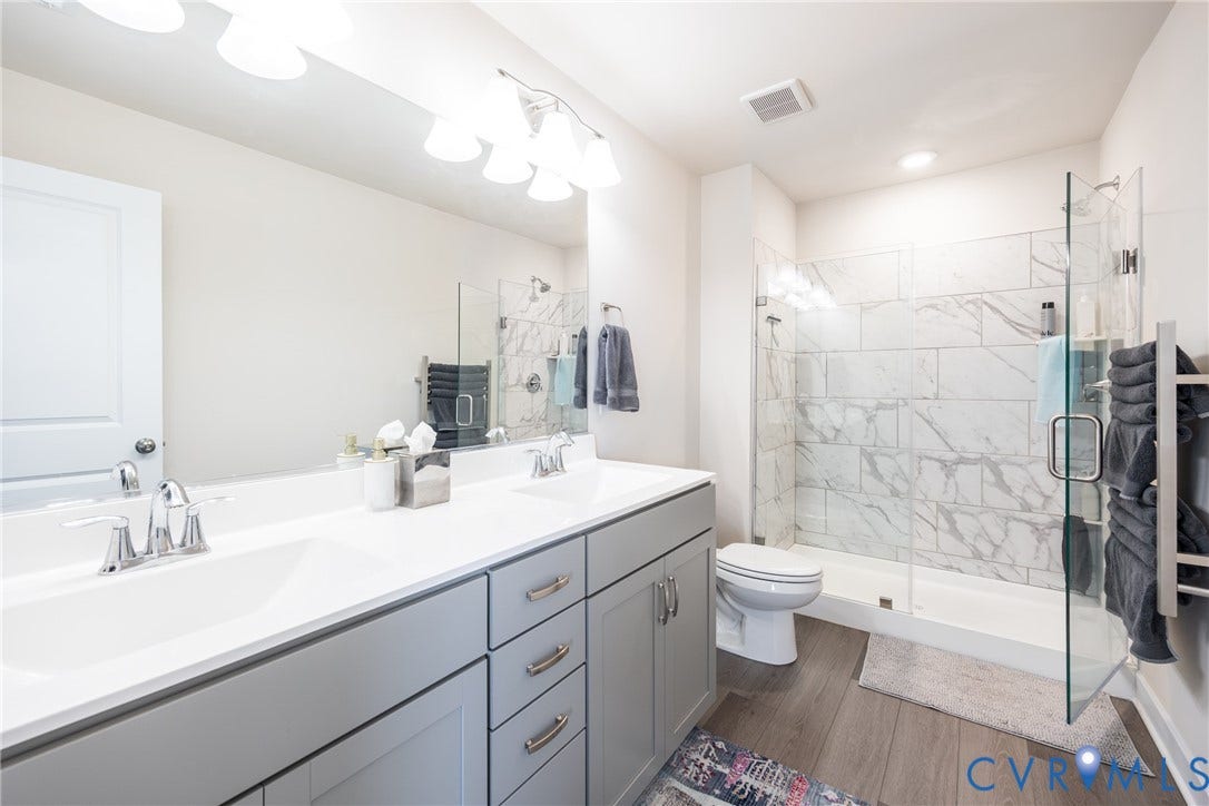12548 Petrel Crossing, Midlothian, VA 23112 Dual-sink vanity with white stone countertop, grey shaker-style cabinetry, walk-in shower with floor-to-ceiling tile surround, glass enclosure, and wood-finish flooring