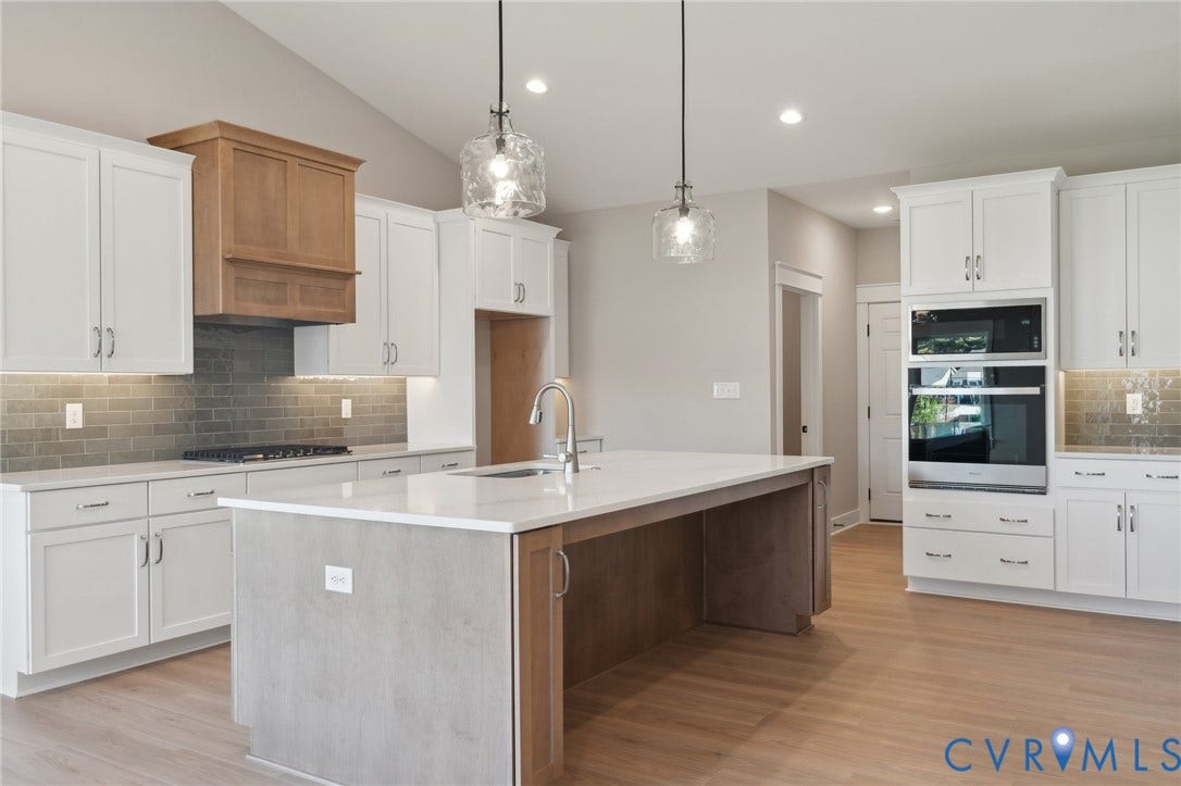 9131 Terroir Lane, Viniterra Villas, New Kent, VA 23124 Kitchen featuring white shaker-style cabinetry, quartz countertops, a central island with a contrasting wood-tone base, integrated stainless steel double wall ovens, and a tile backsplash