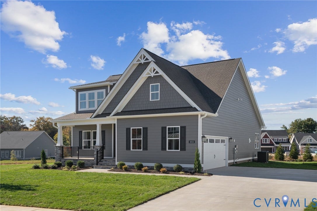 9131 Terroir Lane, Viniterra Villas, New Kent, VA 23124 Two-story residence featuring grey lap siding, stone veneer accents, decorative gable truss detailing, a covered front porch with metal railing, and an attached two-car garage