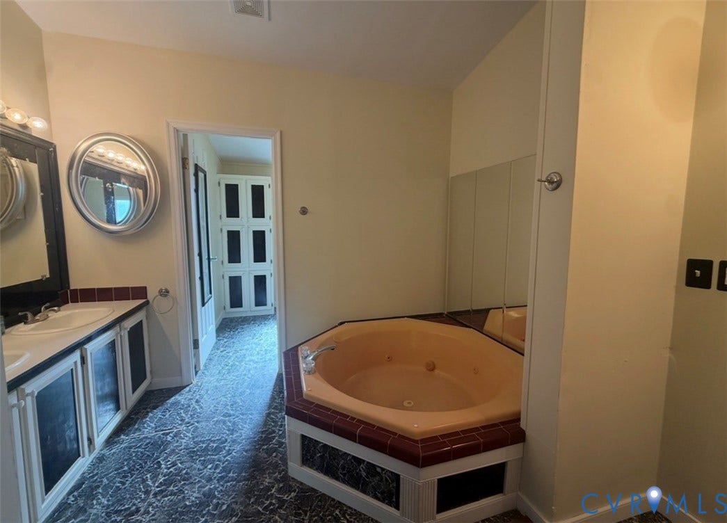 26691 Bell Road, Amelia, VA 23002 Bathroom featuring a dual-sink vanity with dark-toned cabinetry, circular wall mirrors, a drop-in jetted tub with tiled surround, and floor-to-ceiling cabinetry