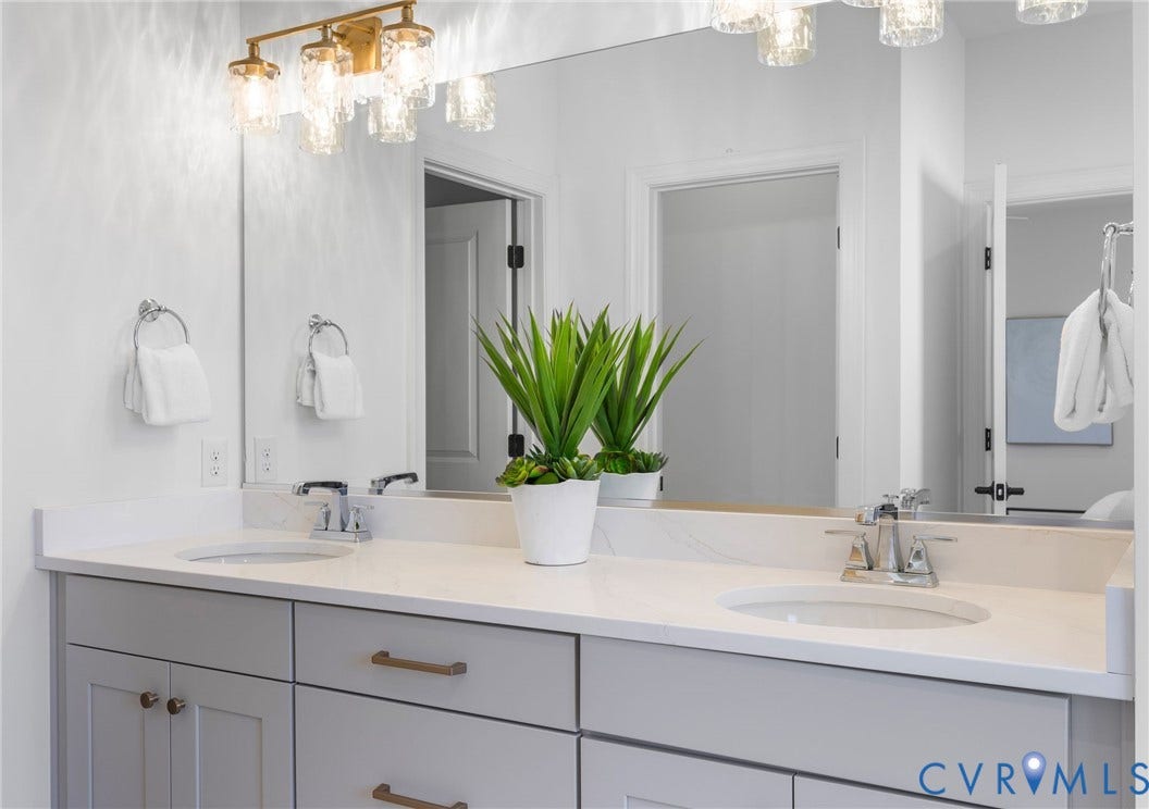 2851 Old Pump Road, Preserve At John Rolfe, Henrico, VA 23233 Dual-sink vanity with a solid-surface countertop, light gray shaker-style cabinetry, metallic hardware, and a wall-to-wall mirror illuminated by multi-light glass fixtures