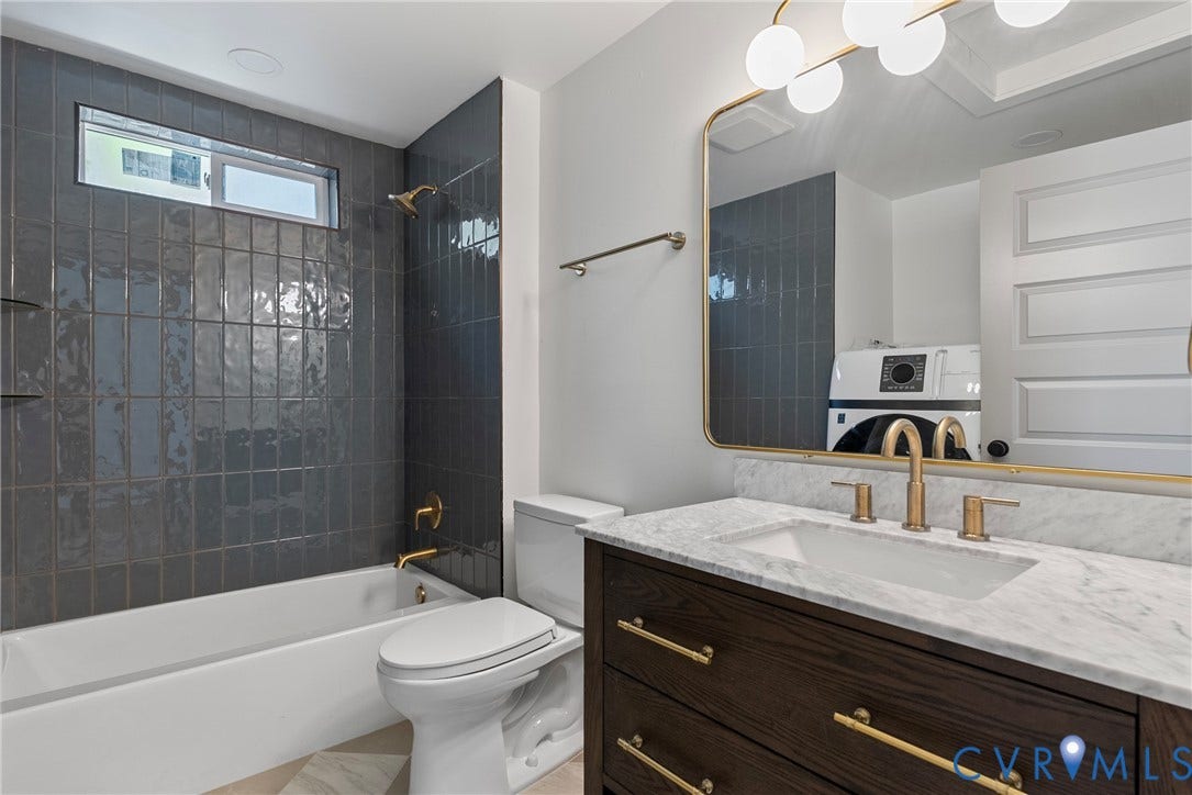 1708 Decatur Street, Swansboro, Richmond, VA 23224 Dark wood vanity with a stone countertop, brass hardware, oversized gold-framed mirror, and vertical charcoal tile bathtub surround