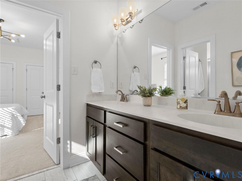 1412 Ascot Hill Terrace, Midlothian, VA 23112 Dark wood cabinetry featuring shaker-style doors, stone-surface countertop with dual undermount sinks, bronze-tone finish faucets, and a large-format vanity mirror with integrated three-bulb light fixture