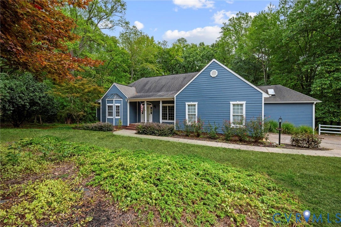 2654 Academy Road, Powhatan, VA 23139 Blue siding exterior featuring a gable roofline, multi-pane windows, a recessed front entrance with a covered porch, and landscaped grounds with mature trees. Virtually edited grass