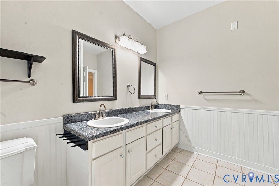 2654 Academy Road, Powhatan, VA 23139 Dual-sink vanity featuring stone-finish countertops, white cabinetry with integrated storage, beadboard wainscoting, and a four-light vanity fixture
