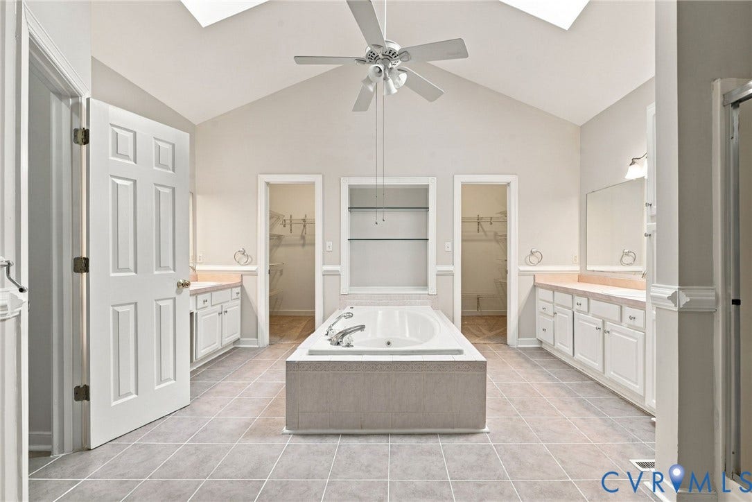 2654 Academy Road, Powhatan, VA 23139 Bathroom featuring a vaulted ceiling with skylights, a central garden tub with tile surround, dual separate white cabinetry vanities, and built-in shelving