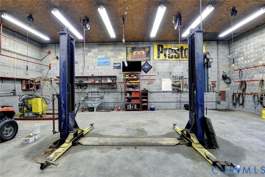 14151 Black Horse Lane, Lanexa, VA 23089 Industrial-style workshop featuring a concrete floor, two-post vehicle lift, exposed concrete block walls, overhead fluorescent lighting, and wall-mounted storage shelving