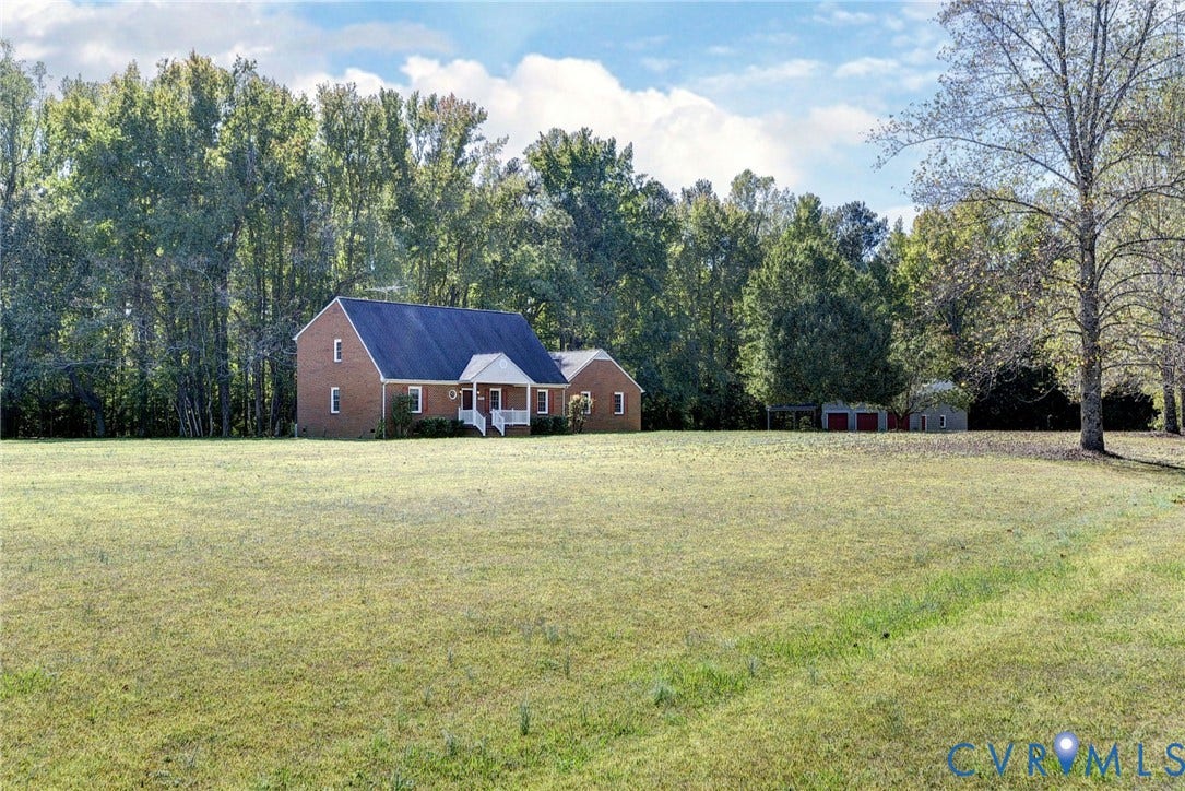 14151 Black Horse Lane, Lanexa, VA 23089 Multi-story brick exterior residence featuring a covered entryway, steeply pitched roof, expansive open lawn, and detached garage structure set against a backdrop of mature trees