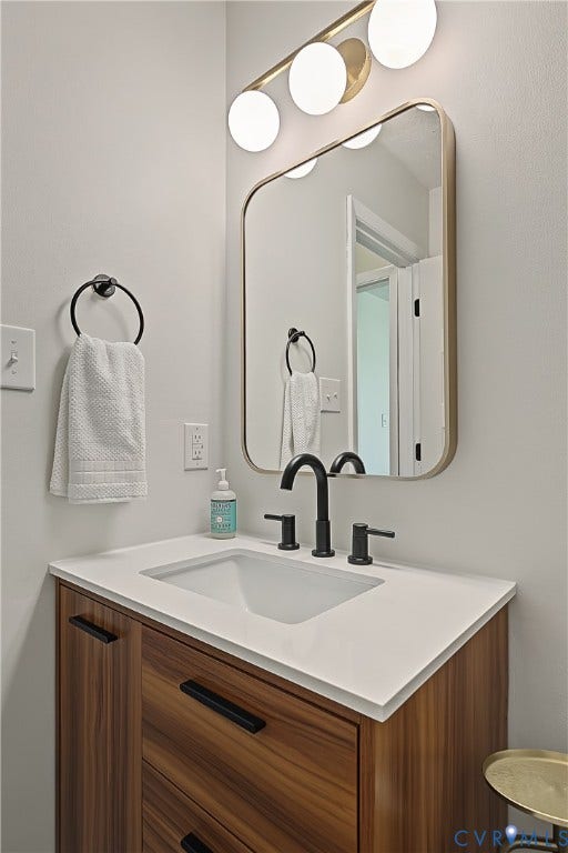 3800 Pheasant Hollow Drive, Henrico, VA 23231 Vanity featuring wood-finish cabinetry, a white countertop, matte black widespread faucet, rectangular mirror with rounded corners, and a three-globe light fixture