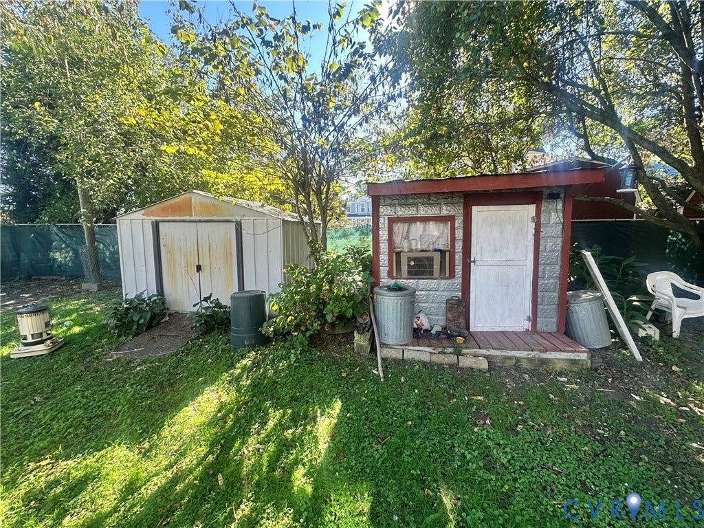5 N Daisy Avenue, Henrico, VA 23075 Rear yard featuring two storage structures, including a metal-sided shed and a frame outbuilding with a porch, surrounded by mature trees