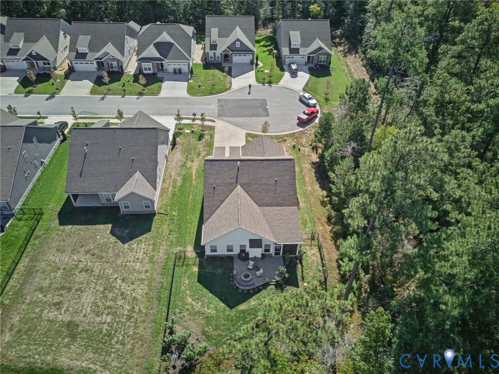 Aerial view of residence featuring a concrete driveway, a rear paver patio with a stone fire pit, and a grass-covered lot bordering a wooded perimeter