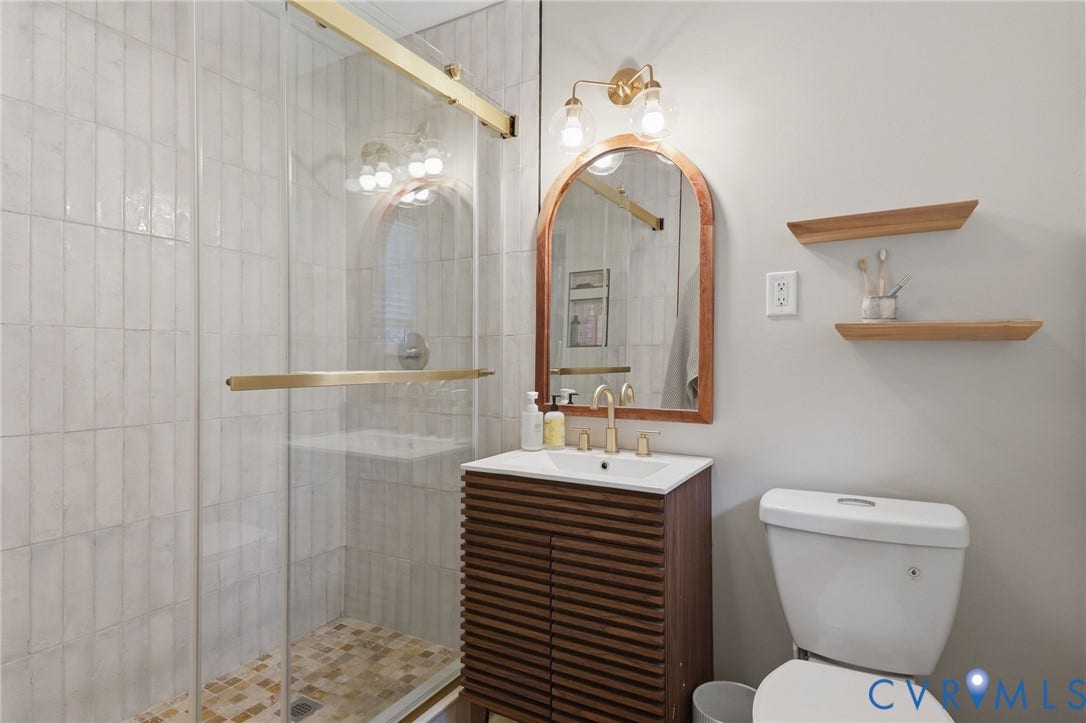 9 E 31st Street, Richmond, VA 23224 Bathroom featuring a slatted wood-finish vanity, arched mirror with brass-tone frame, glass-enclosed shower with subway-style wall tile, and brass-tone plumbing fixtures