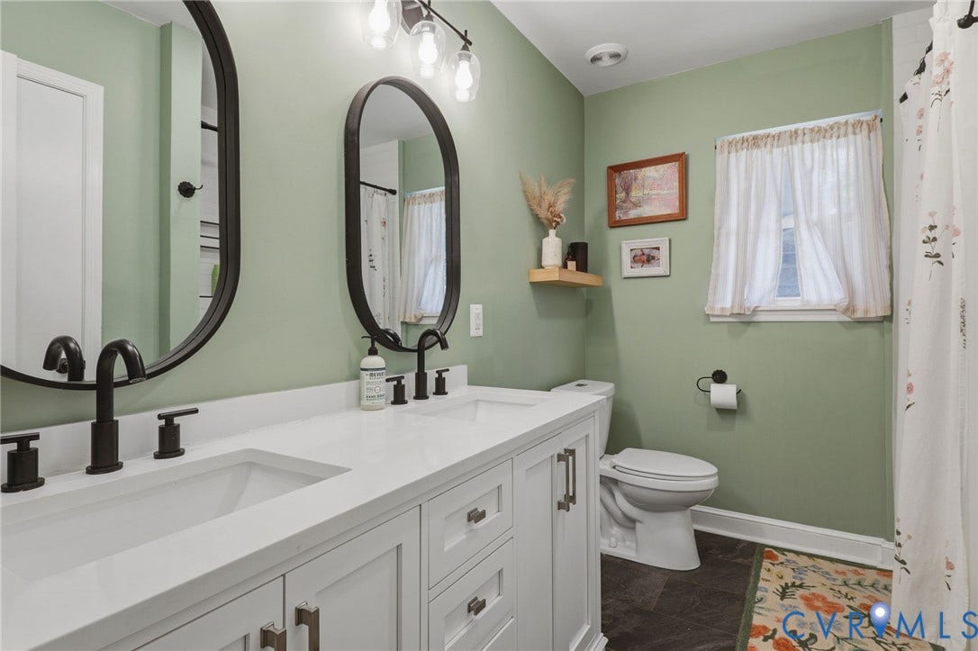 9 E 31st Street, Richmond, VA 23224 Dual-sink vanity with white cabinetry, solid surface countertop, matte black fixtures, oval mirrors, and integrated overhead lighting