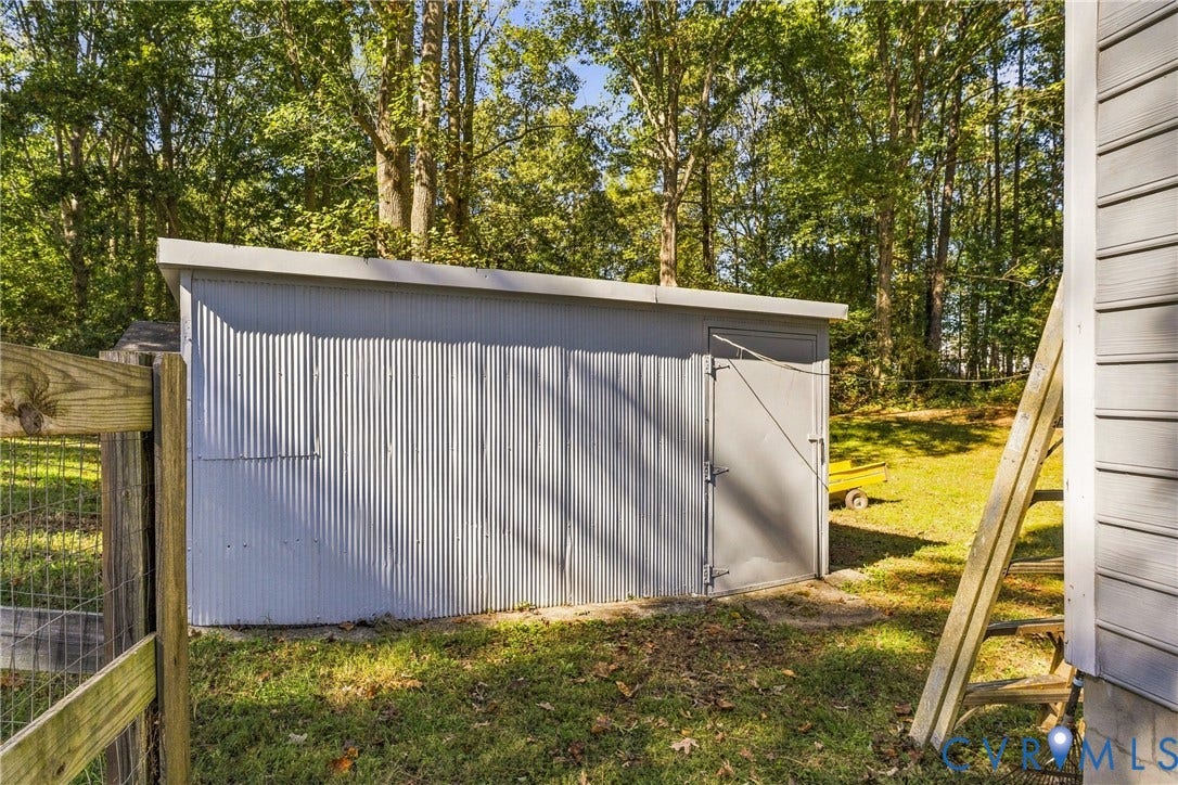 8840 Black Creek Road, New Kent, VA 23141 Exterior storage shed featuring corrugated metal siding, a flat roofline, and a solid swing door, situated on a grass lot with mature perimeter trees