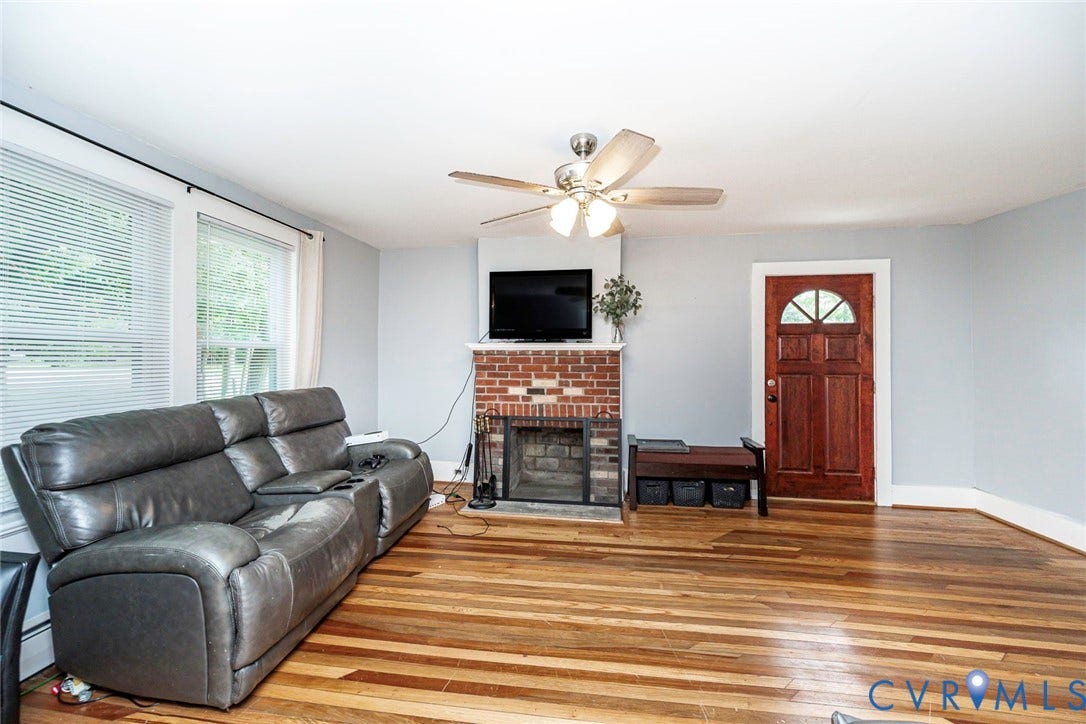 1818 Arlington Road, Hopewell, VA 23860 Living area featuring wood-finish flooring, a red brick fireplace with a built-in mantel, a ceiling fan with light fixture, and a solid wood exterior door with decorative glass paneling
