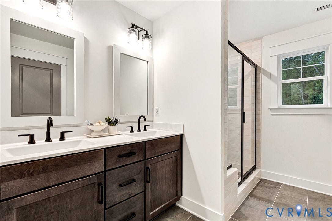 9107 Terroir Lane, Viniterra Villas, New Kent, VA 23124 Dual-sink vanity with dark wood-finish cabinetry, light-toned countertop, matte black hardware, and framed mirrors with overhead industrial-style lighting, complemented by a walk-in shower with black-frame glass enclosure and large-format tile flooring