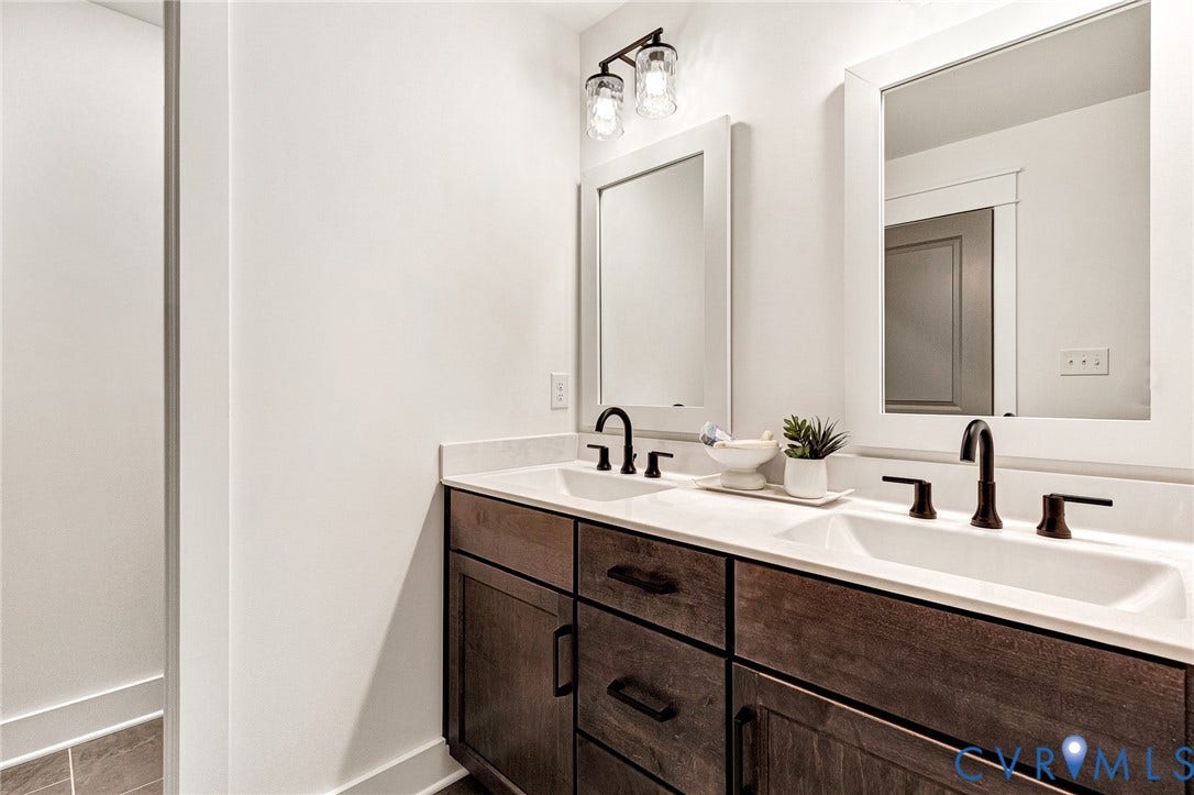 9107 Terroir Lane, Viniterra Villas, New Kent, VA 23124 Dual-sink vanity featuring dark wood-finish cabinetry, a white countertop, rectangular under-mount sinks, and matte black hardware with matching wall-mounted lighting