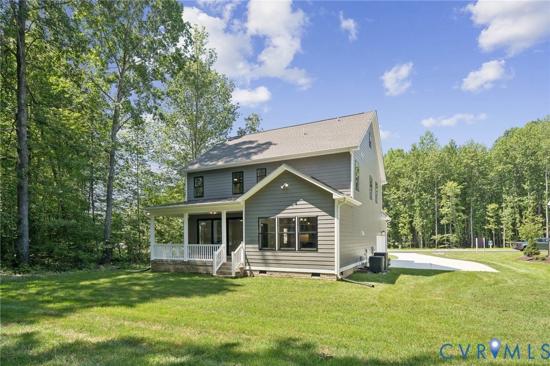 9107 Terroir Lane, Viniterra Villas, New Kent, VA 23124 Two-story residence featuring grey lap siding, a covered porch with white railing, and a concrete driveway, situated on a lot with mature perimeter trees