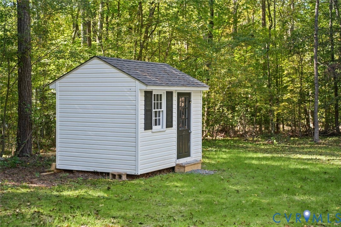 15227 Horseshoe Bridge Road, Hanover, VA 23047 Detached outbuilding featuring white vinyl siding, a gabled roof, dark shutters, and a single-entry door, set within a grassed lot bordered by mature trees