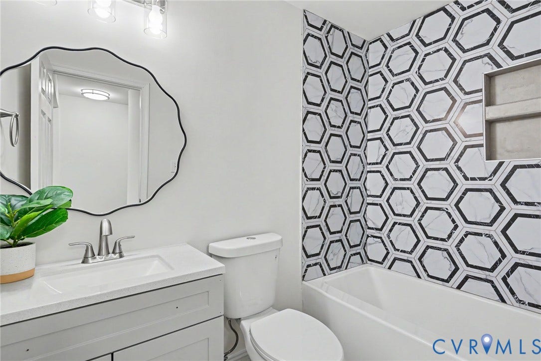 1210 N 24th Street, Church Hill Central, Richmond, VA 23223 Grey vanity with integrated countertop and sink, scalloped-edge mirror, multi-light fixture, and bathtub featuring a hexagonal patterned tile surround with a built-in shower niche
