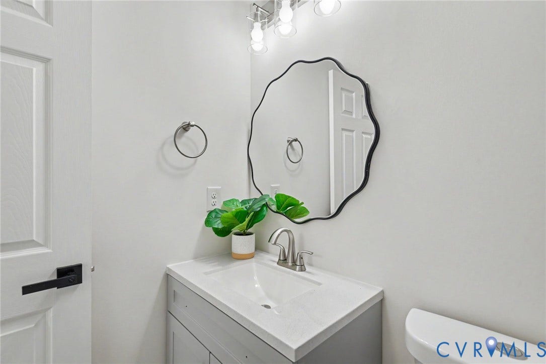 1210 N 24th Street, Church Hill Central, Richmond, VA 23223 Vanity featuring a light-toned countertop, integrated basin, and brushed-nickel dual-handle faucet, accented by a scalloped-edge wall mirror and overhead multi-bulb vanity light