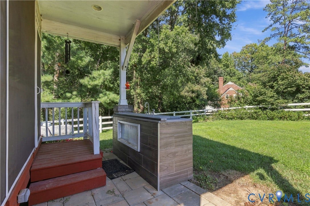 8307 Wallingford Lane, Chickahominy Bluffs, Henrico, VA 23227 Covered patio featuring paver flooring, built-in outdoor kitchen with wood-finish tile cladding, and wood-railing stairs, overlooking a manicured lawn enclosed by a white fence