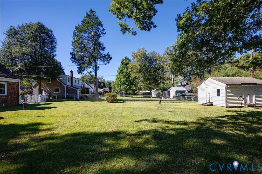 Expansive backyard featuring a large grass lawn, mature trees, chain-link perimeter fencing, and a detached storage structure with vinyl siding