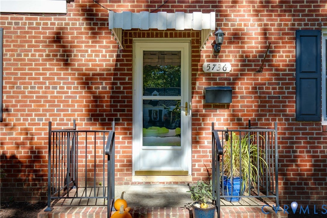 Red brick exterior featuring a white storm door, scalloped metal awning, dark-toned shutters, and a black wall-mounted mailbox