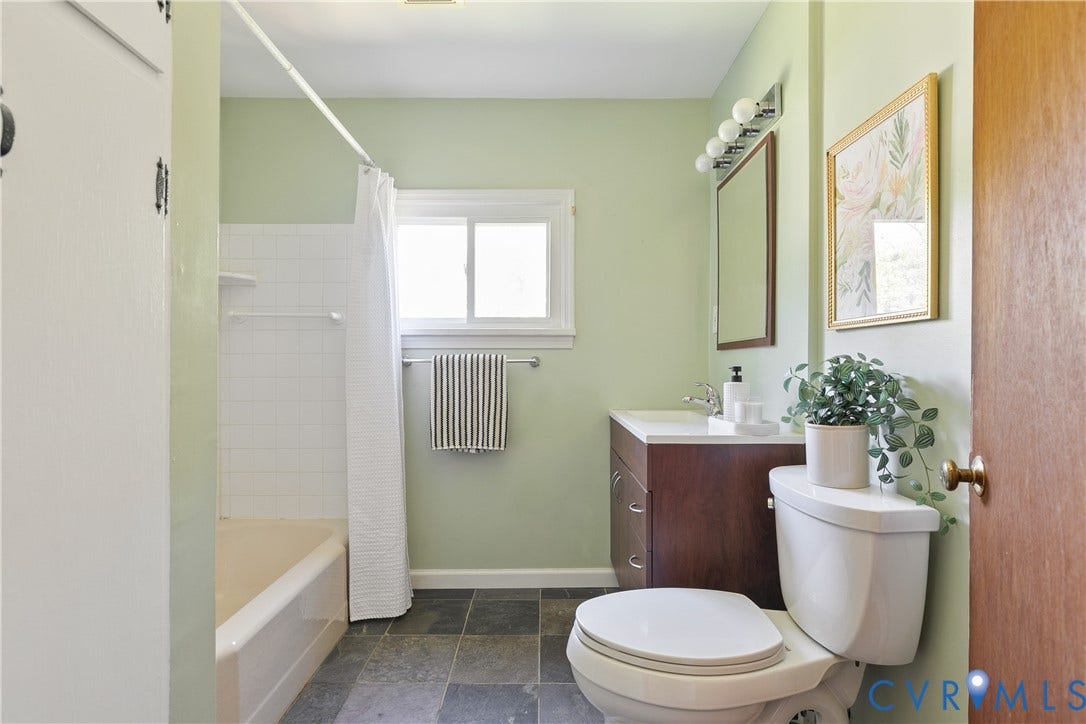 Bathroom featuring natural stone tile flooring, a wood-finish vanity with a rectangular sink, a wall-mounted vanity mirror with a multi-bulb light fixture, and a white tub-shower combination with tile surround