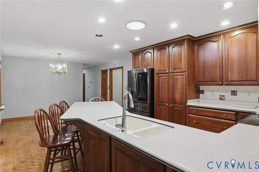 Kitchen featuring rich wood-finish cabinetry, a central island with a dual-basin sink, high-reach faucet, neutral solid-surface countertops, and recessed lighting