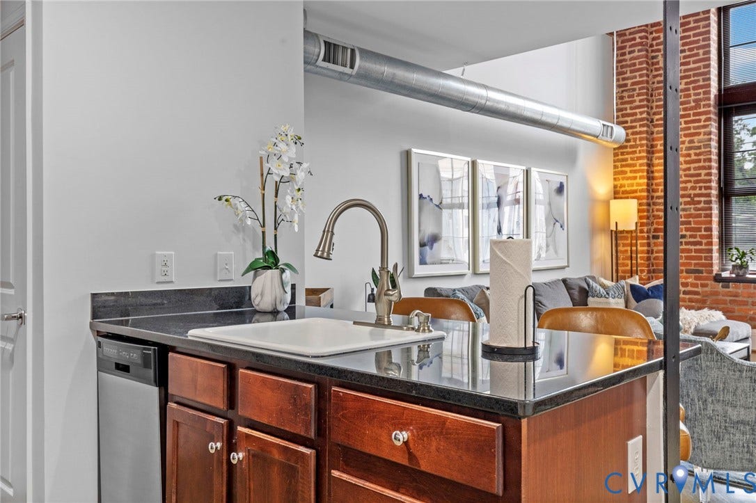 815 Porter Street, #U301, Richmond, VA 23224 Kitchen island featuring dark stone countertops, an under-mount square sink, gooseneck faucet, warm wood-finish cabinetry, and exposed brick walls