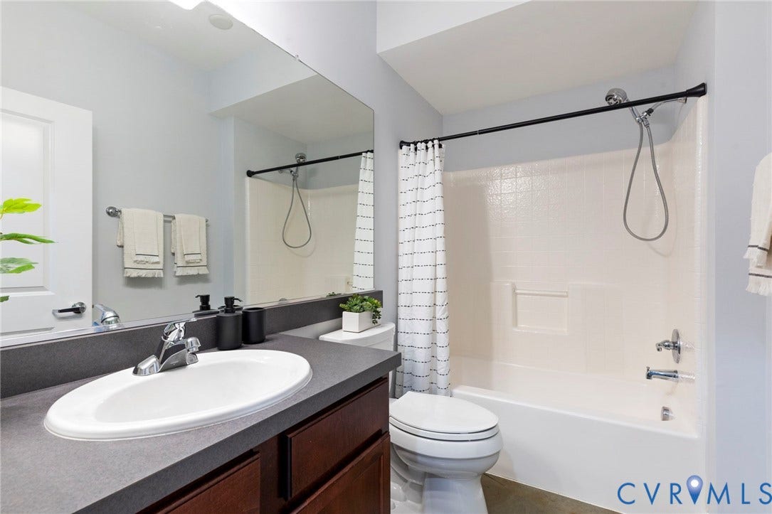 815 Porter Street, #U301, Richmond, VA 23224 Full-width vanity with a dark countertop and integrated sink, complemented by a large wall-mounted mirror, wood-finish cabinetry, and a shower-tub combination with a handheld fixture