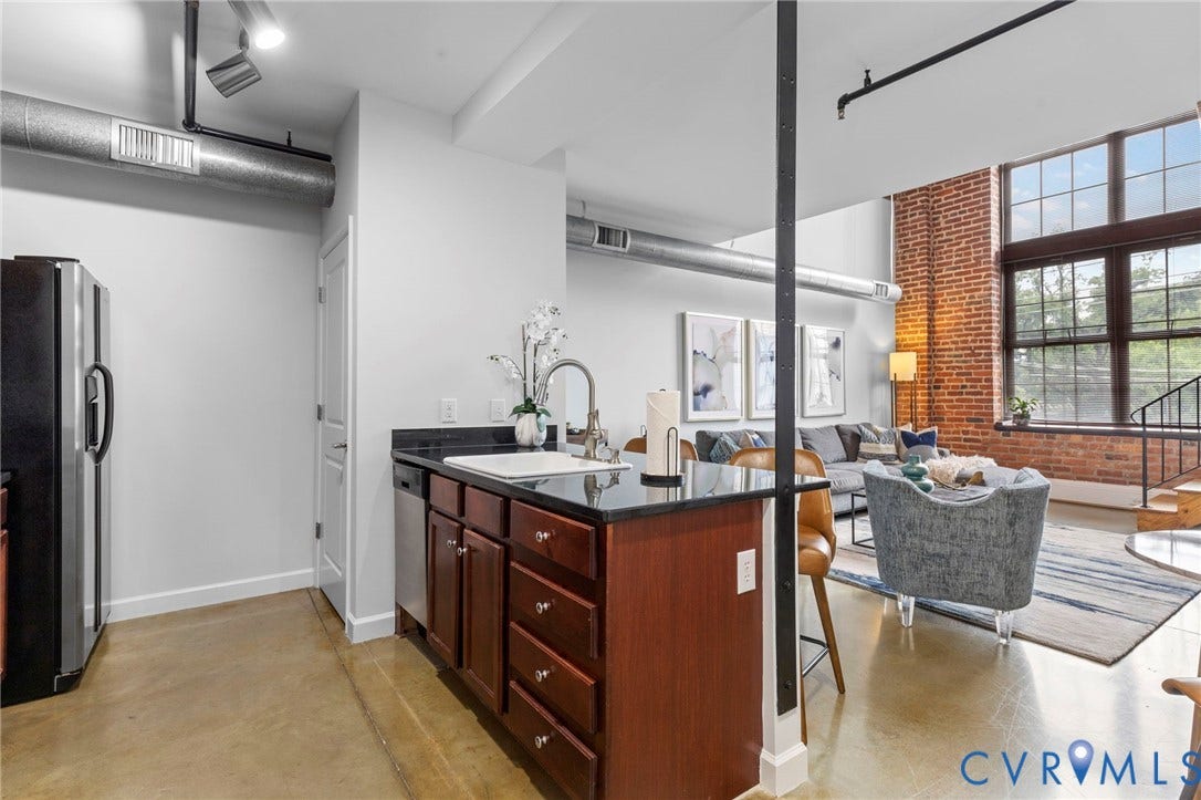 815 Porter Street, #U301, Richmond, VA 23224 Industrial-style loft featuring exposed brick walls, expansive multi-pane windows, polished concrete flooring, open-concept kitchen with dark stone countertops, and overhead structural steel support