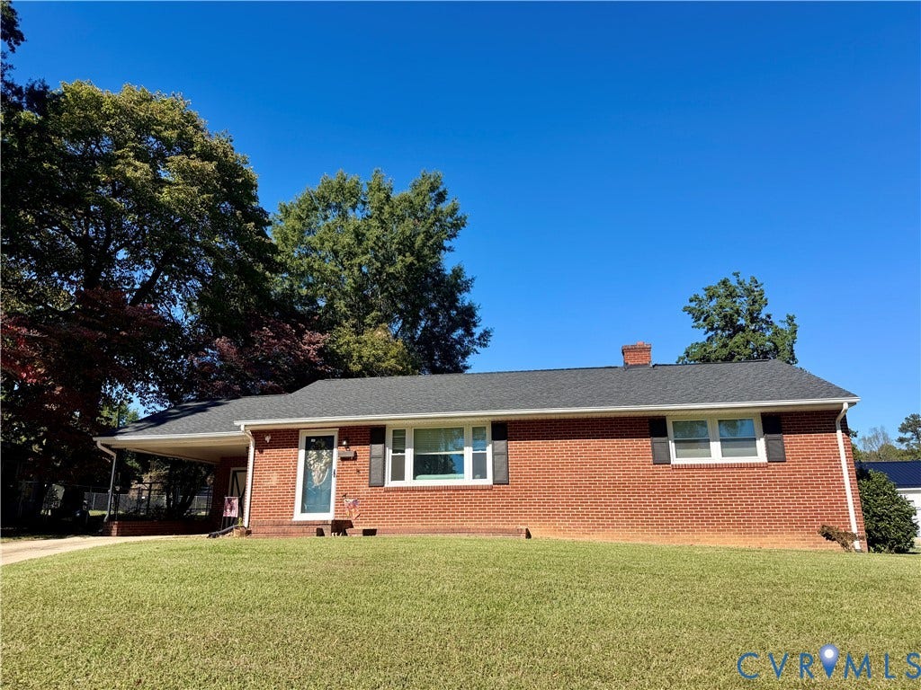 Red brick exterior featuring a covered carport, asphalt shingle roof, dark shutters, and mature trees