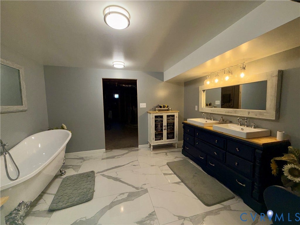 Freestanding clawfoot tub with hand-held shower attachment, dual-sink vanity with dark-finish cabinetry, large-format veined tile flooring, and multi-light vanity fixtures