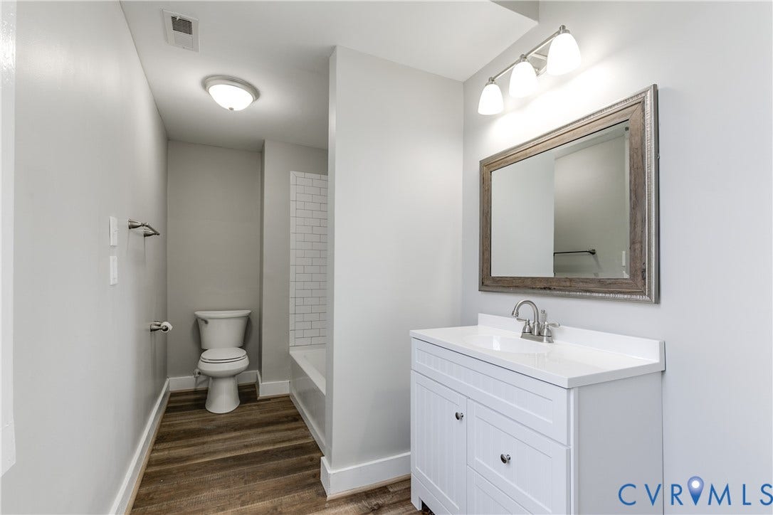 620 Chimborazo Boulevard, Church Hill, Richmond, VA 23223 White vanity cabinet featuring a solid surface countertop, framed mirror, three-light fixture, wood-finish flooring, and tub surround with white subway tile