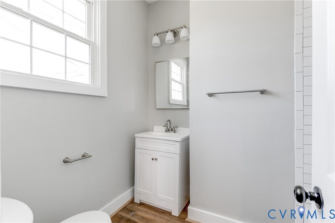 620 Chimborazo Boulevard, Church Hill, Richmond, VA 23223 White vanity with a neutral solid surface countertop, rectangular under-mount sink, three-light polished chrome vanity fixture, framed wall mirror, and wood-finish flooring