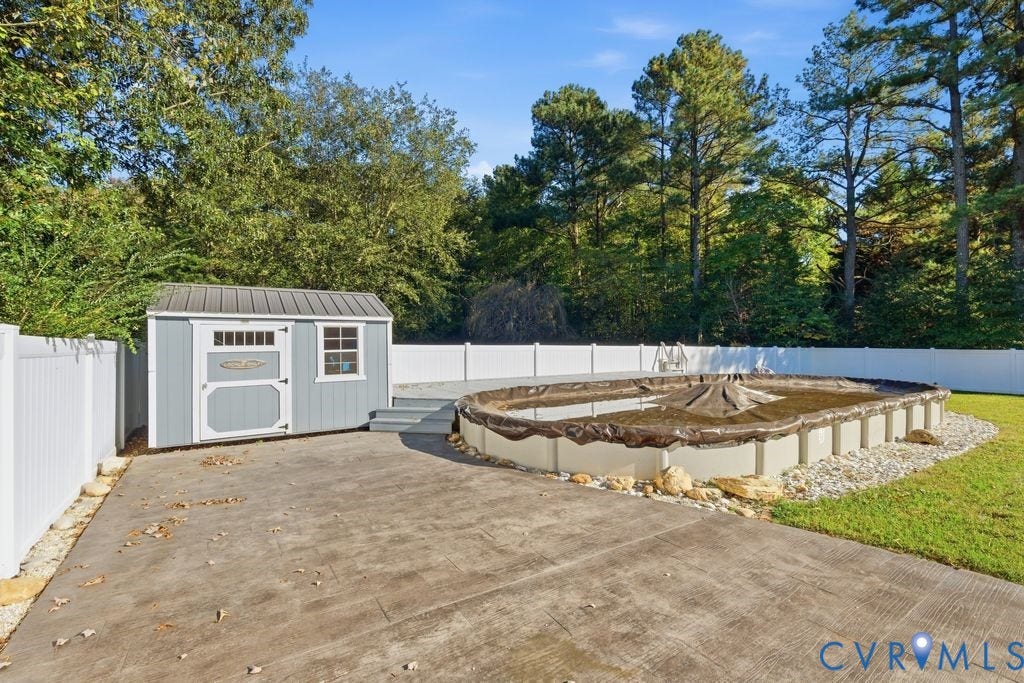 8477 Cheraw Road, Mechanicsville, VA 23116 Backyard featuring an above-ground pool, concrete patio, detached storage structure with a metal roof, and white vinyl perimeter fencing