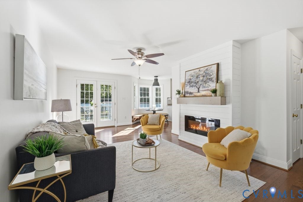 8477 Cheraw Road, Mechanicsville, VA 23116 Spacious interior featuring a white shiplap fireplace surround, wooden mantel, wood-finish flooring, double glass-paneled exterior doors, and a ceiling fan